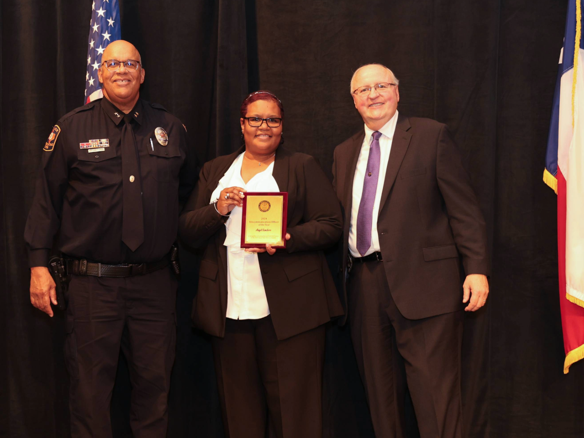Uniformed officer, award recipient holding a plaque, and suited administrator pose on stage between U.S. and Texas flags.